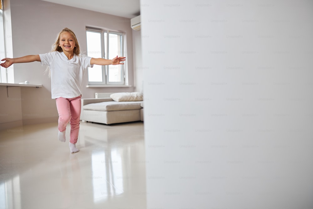 Horizontal image of happy funny little girl in stylish casual clothes running in the big apartment to meet