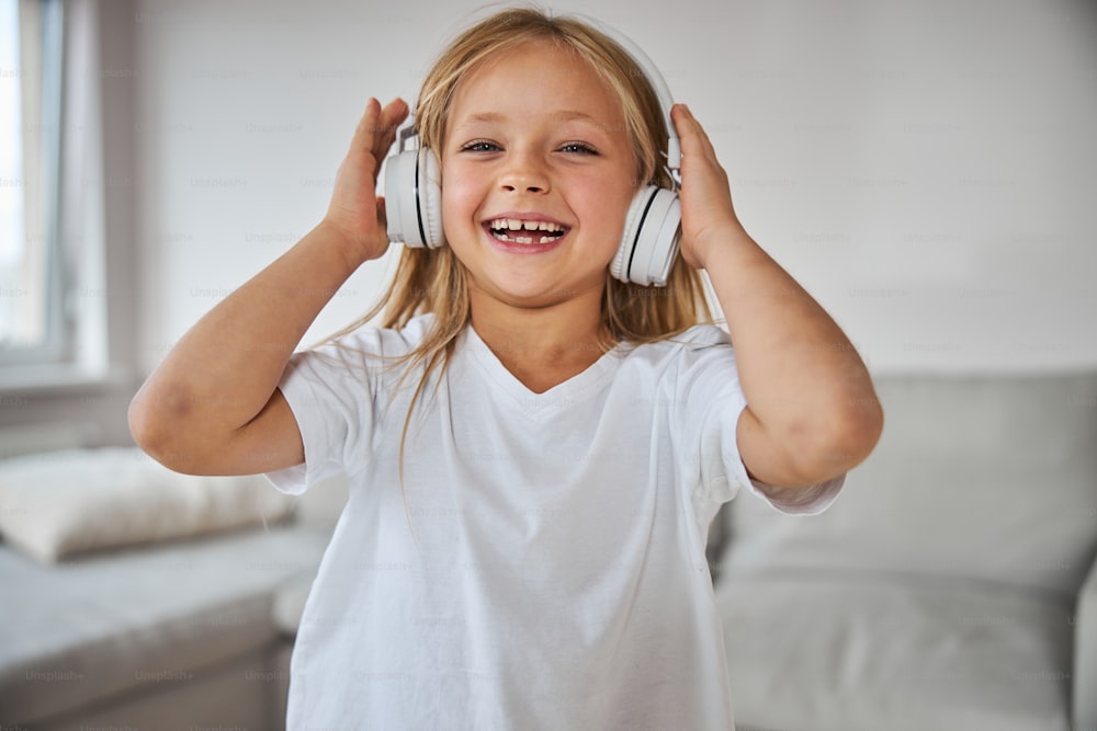Close up portrait of happy cheerful little girl in white shirt listening something in headphones and posing at the camera in white room