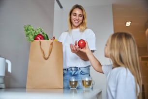 Side view portrait of small blonde daughter taking fruit from her mother while sitting at the white table in room inside