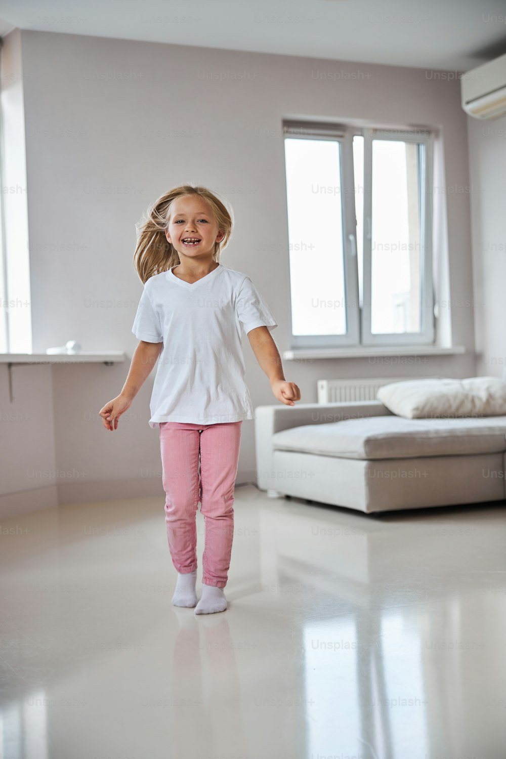 Retrato de cuerpo entero de una niña pequeña sonriente feliz que se divierte en la habitación interior mientras está de pie frente al sofá gris
