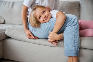 Close up portrait of cute elegant little girl spending time with her lovely kind mother while sitting on the grey sofa in room inside