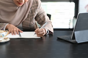 Ausschnittaufnahme einer glücklichen muslimischen Geschäftsfrau im Hijab nimmt Notiz neben Computer-Tablet auf dem Tisch im Büro.