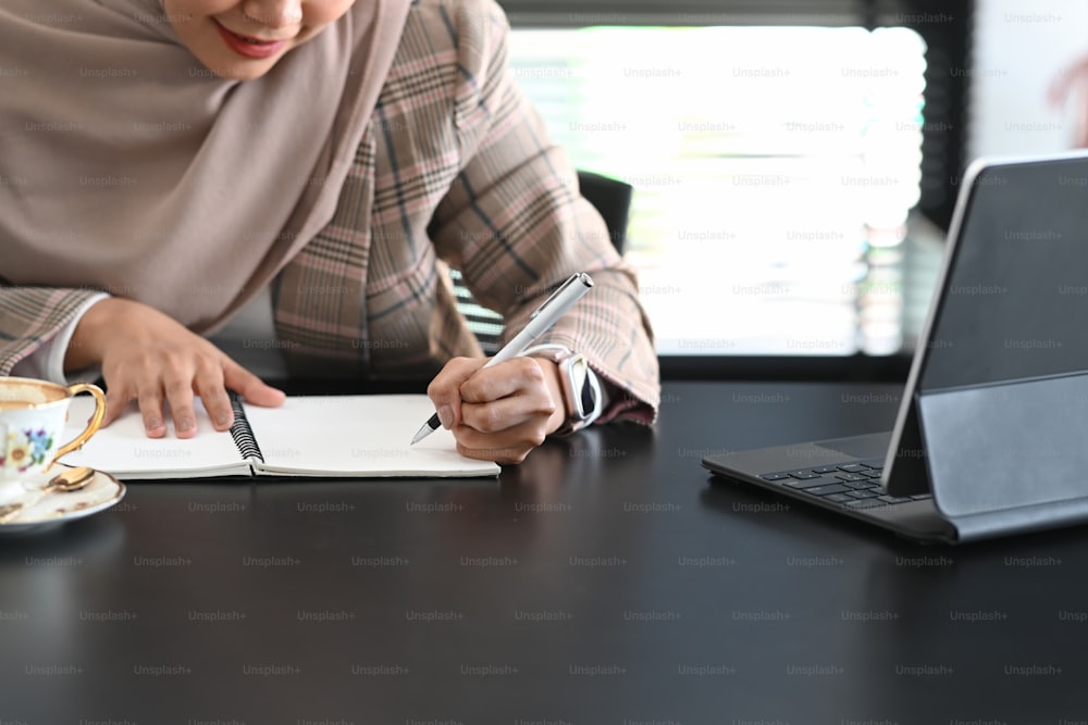 Cropped shot of happy muslim businesswoman in hijab is taking note beside computer tablet on table in office.