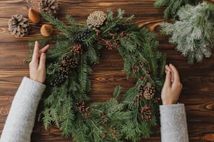 Mãos de florista segurando coroa de Natal rústica em mesa de madeira rústica com bagas, pinhas, decorações festivas naturais, flat lay. Workshop sazonal de inverno, advento de férias