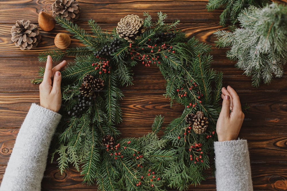 Florist hands holding rustic christmas wreath on rustic wooden table with berries, pine cones, natural festive decorations, flat lay. Seasonal winter workshop, holiday advent