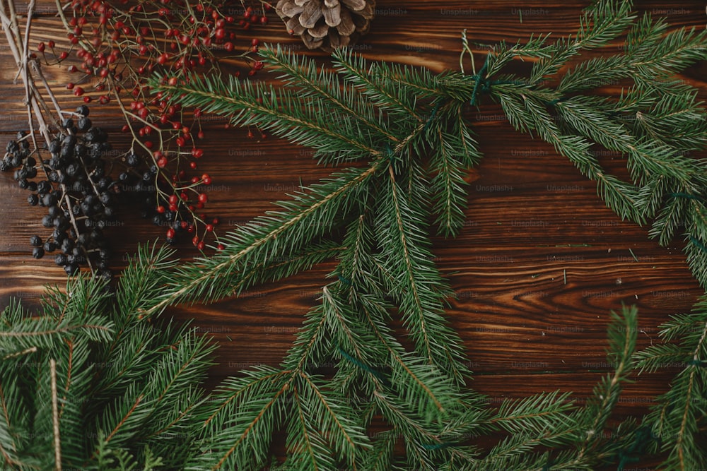 나무 테이블에 소박한 크리스마스 화환을 만듭니다. 빨간 열매, 녹색 전나무 가지, 솔방울, 소박한 배경 테이블에 자연 축제 장식이 있는 크리스마스 화환