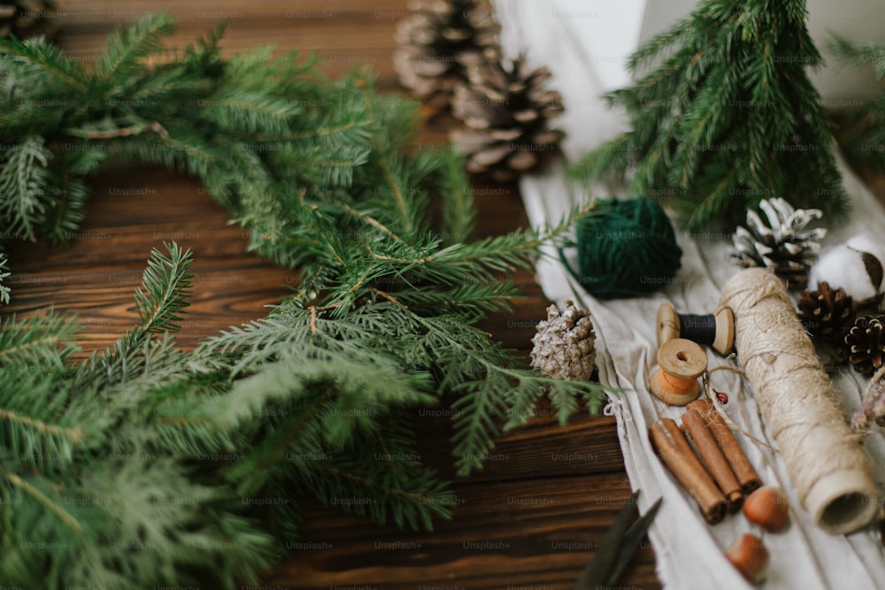 Weihnachtskranz mit grünen Zweigen, Tannenzapfen und natürlichen festlichen Dekorationen, Scheren, Bindfäden. Uriger Weihnachtskranz auf Holztisch basteln, saisonaler Feiertagsadvent