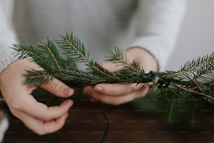 Close up of female hands holding green fir branches and making rustic christmas wreath on background of wooden table. Handmade festive decor, holiday advent. Seasonal winter workshop