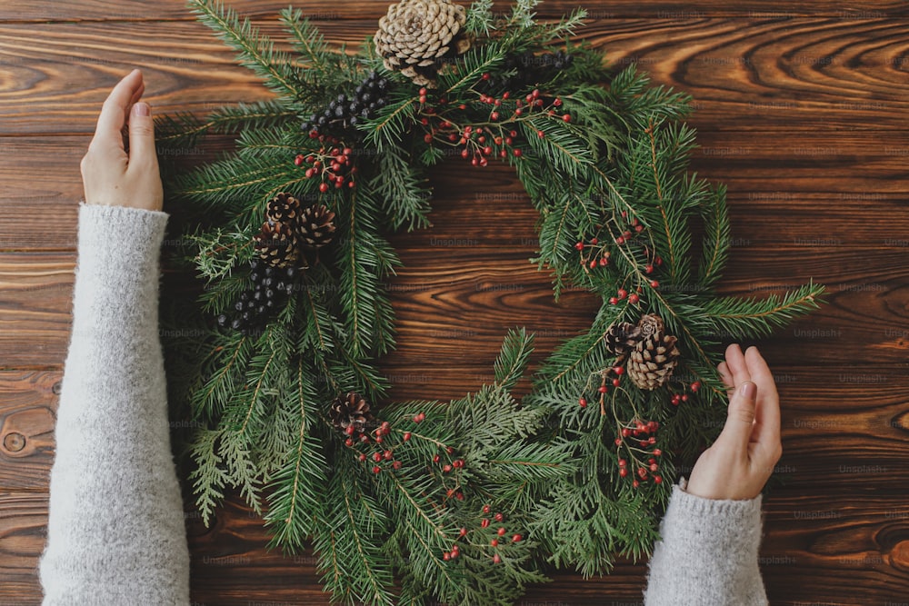 Ghirlanda di natale rustica a posa piatta. Mani del fiorista che tengono rami di abete verde su tavolo di legno rustico con bacche, pigne, decorazioni festive naturali. Laboratorio invernale stagionale, festa dell'Avvento