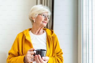 Ältere Frau am Fenster mit einer Tasse Kaffee oder Tee. Ältere Frau isoliert, um eine Infektion zu verhindern. Alte Frau schaut durch das Fenster