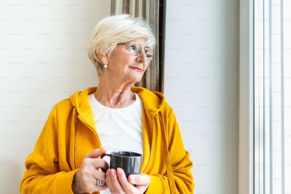 Mujer mayor en la ventana sosteniendo una taza de café o té. Mujer anciana aislada para prevenir contagios. Anciana mirando por la ventana
