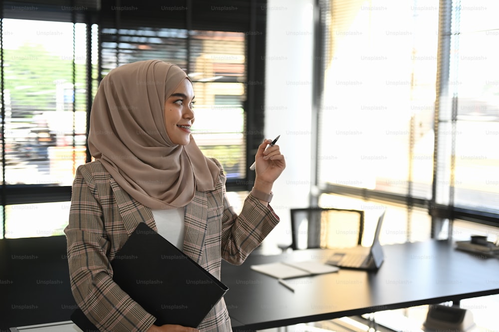 Uma mulher muçulmana alegre em abaya de pé no escritório segurando o arquivo de documentos enquanto olhava pela janela.