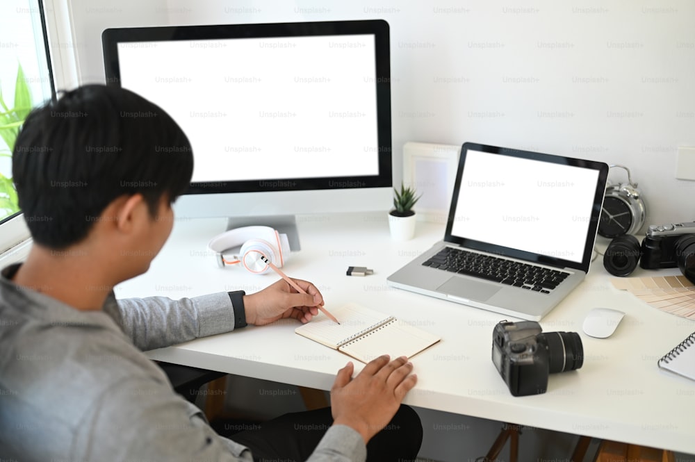 Un graphiste ou un photographe prend des notes alors qu’il est assis devant plusieurs ordinateurs sur son lieu de travail.