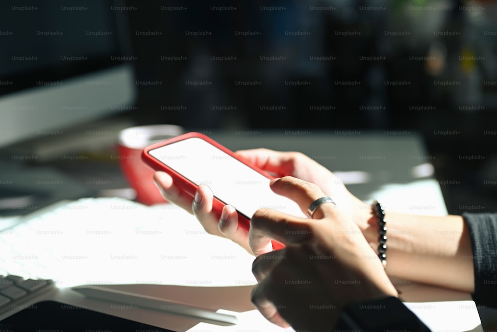 Vista de cerca de la escritura femenina en la pantalla en blanco de la maqueta del teléfono móvil mientras está sentada en el espacio de trabajo. Teléfono móvil con pantalla en blanco para montaje gráfico.