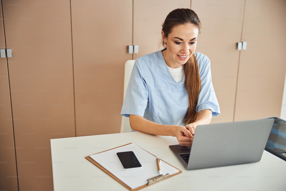 Signora bruna sorridente in uniforme da medico seduta al tavolo e guardando lo schermo del computer portatile