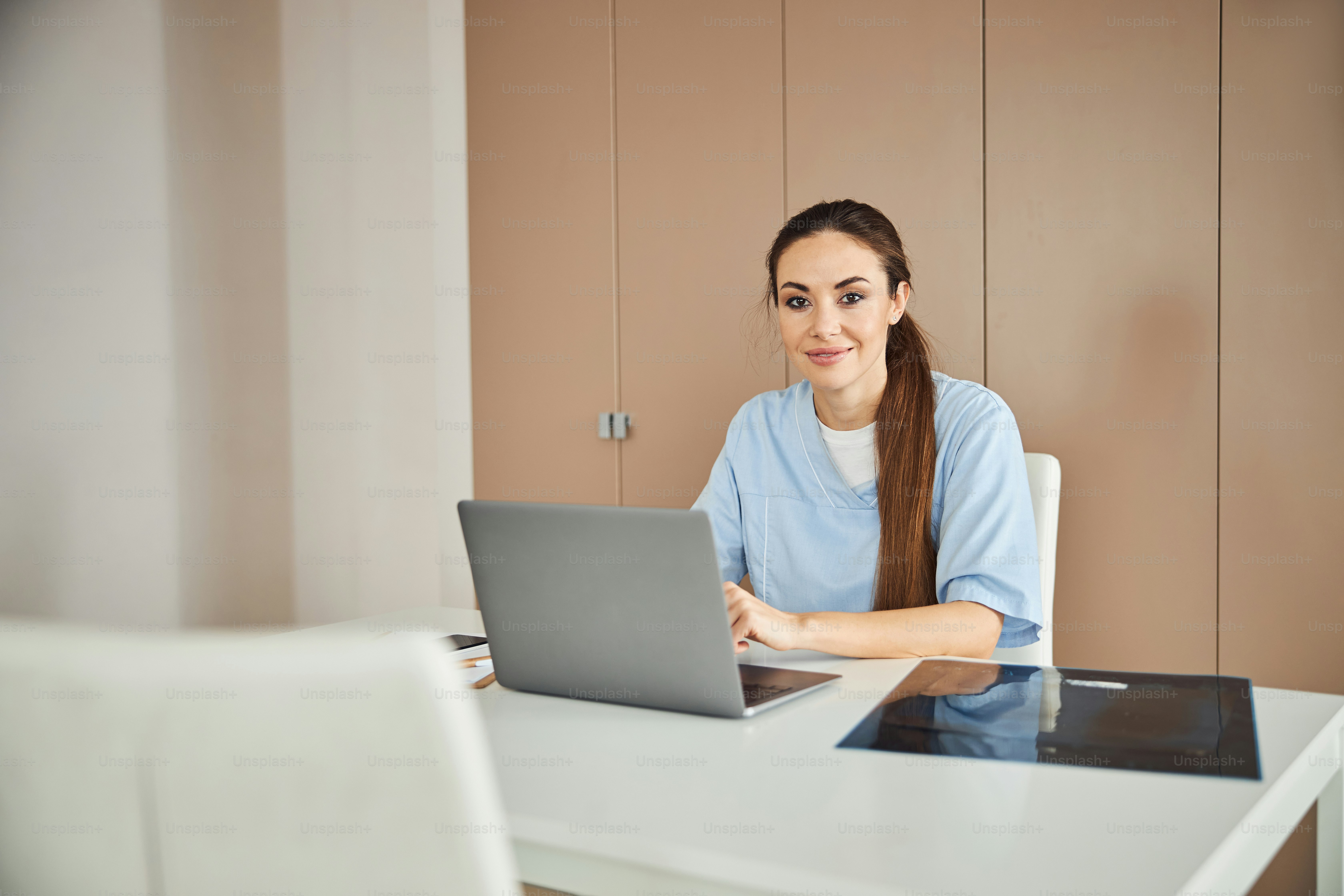 Pretty brunette lady in medical scrubs sitting at the table with laptop and looking at the camera