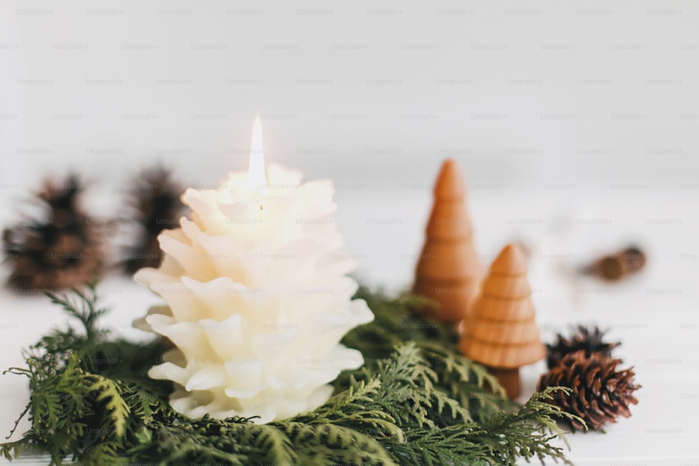 Merry Christmas! Stylish pine cone candle, cedar wreath and little wooden christmas trees on rustic white table. Festive modern decor, holiday table setting