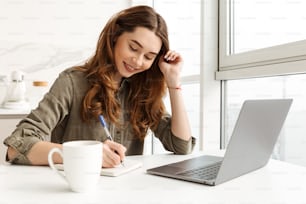 Smart business woman sitting in kitchen at home and writing down notes while working on notebook