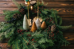 Merry Christmas! Modern handmade wreath with little trees, candles and pine cones on rustic wooden table, christmas table setting. Festive stylish decor for holiday dinner at home.