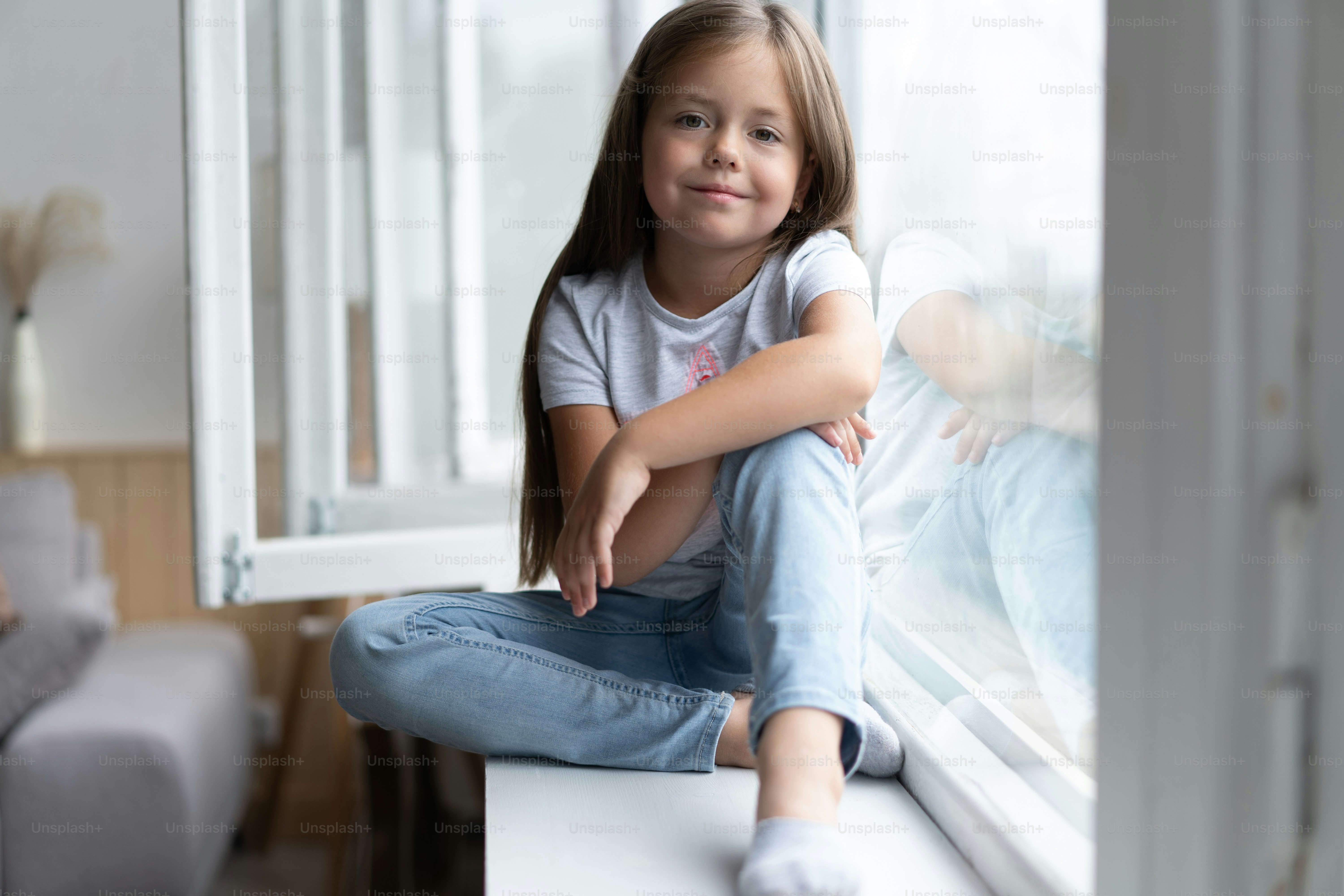 Beautiful little girl smiling and watching out the window. A child looks out the window. Young girl looking from window. Portrait of cheerful kid lies at windowsill.