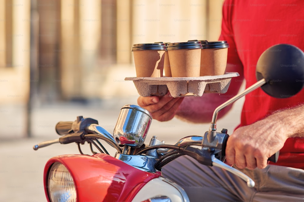 Mensageiro masculino entregando quatro xícaras de café em scooter, de pé em uma rua ensolarada. Conceito de entrega de alimentos