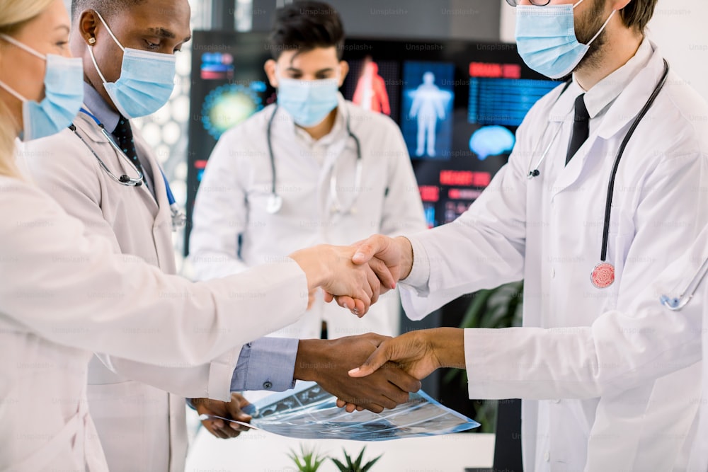 Pandemy, outbreak of coronavirus concept. Close up cropped image of multiethnic group of doctors with face masks, shaking hands at the meeting. Focus on hands of African and Caucasian colleagues.