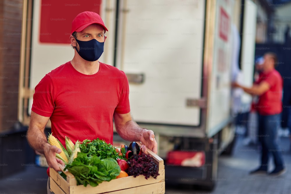 Stay home. Young caucasian male courier wearing black face protective mask holding grocery box, standing on the street with truck on the background. Food delivery during covid 19 outbreak concept