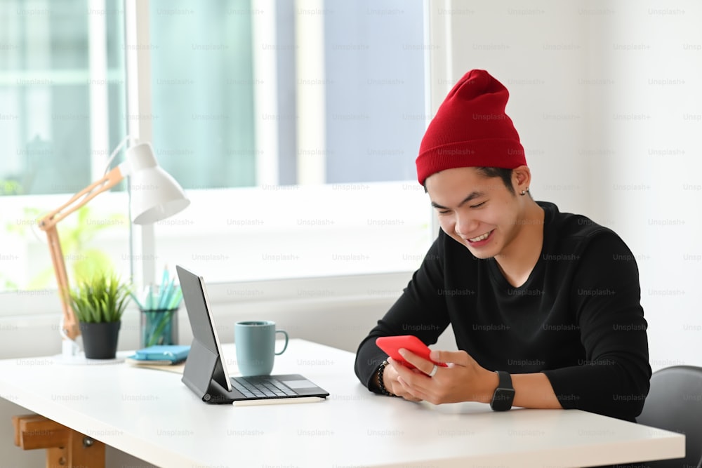 陽気で魅力的な若い男性のグラフィックデザイナーは、笑顔で、モダンなオフィスの机でスマートフォンを使用しています。