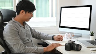 Side view of graphic designer or photographer is sitting in front of empty screen computer and taking notes in notebook.