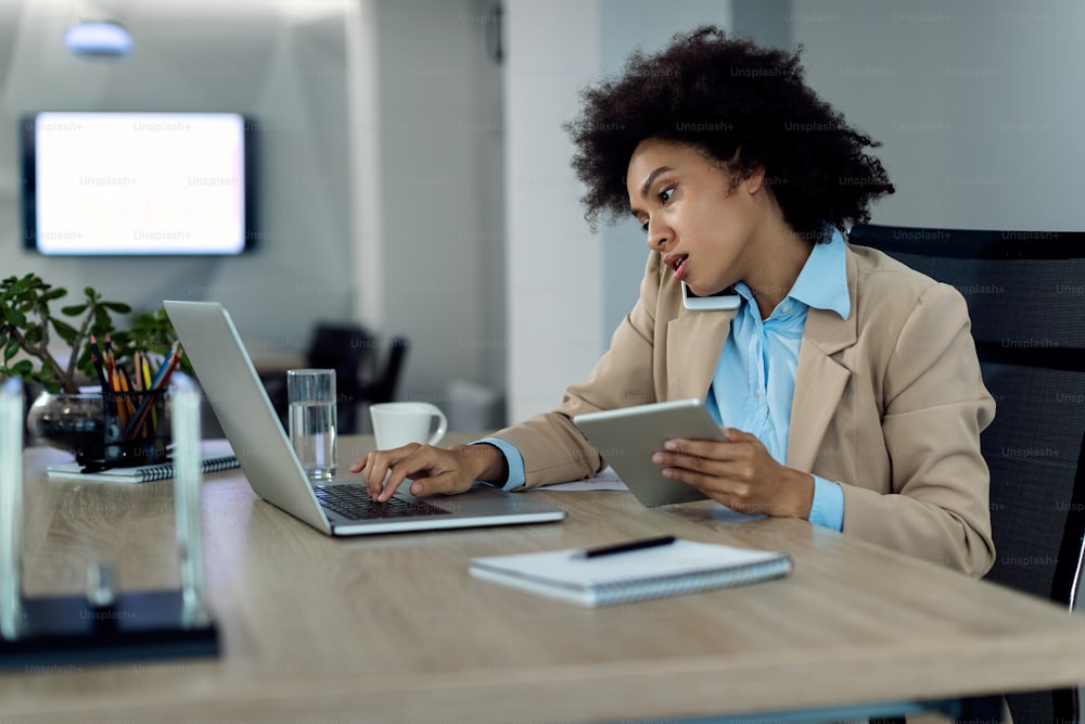 Multitasking-schwarze Geschäftsfrau, die am Laptop arbeitet, während sie das Touchpad benutzt und im Büro telefoniert.