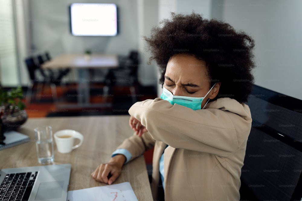 Empresaria negra tosiendo en el codo mientras usa una máscara facial protectora y trabaja en la oficina.