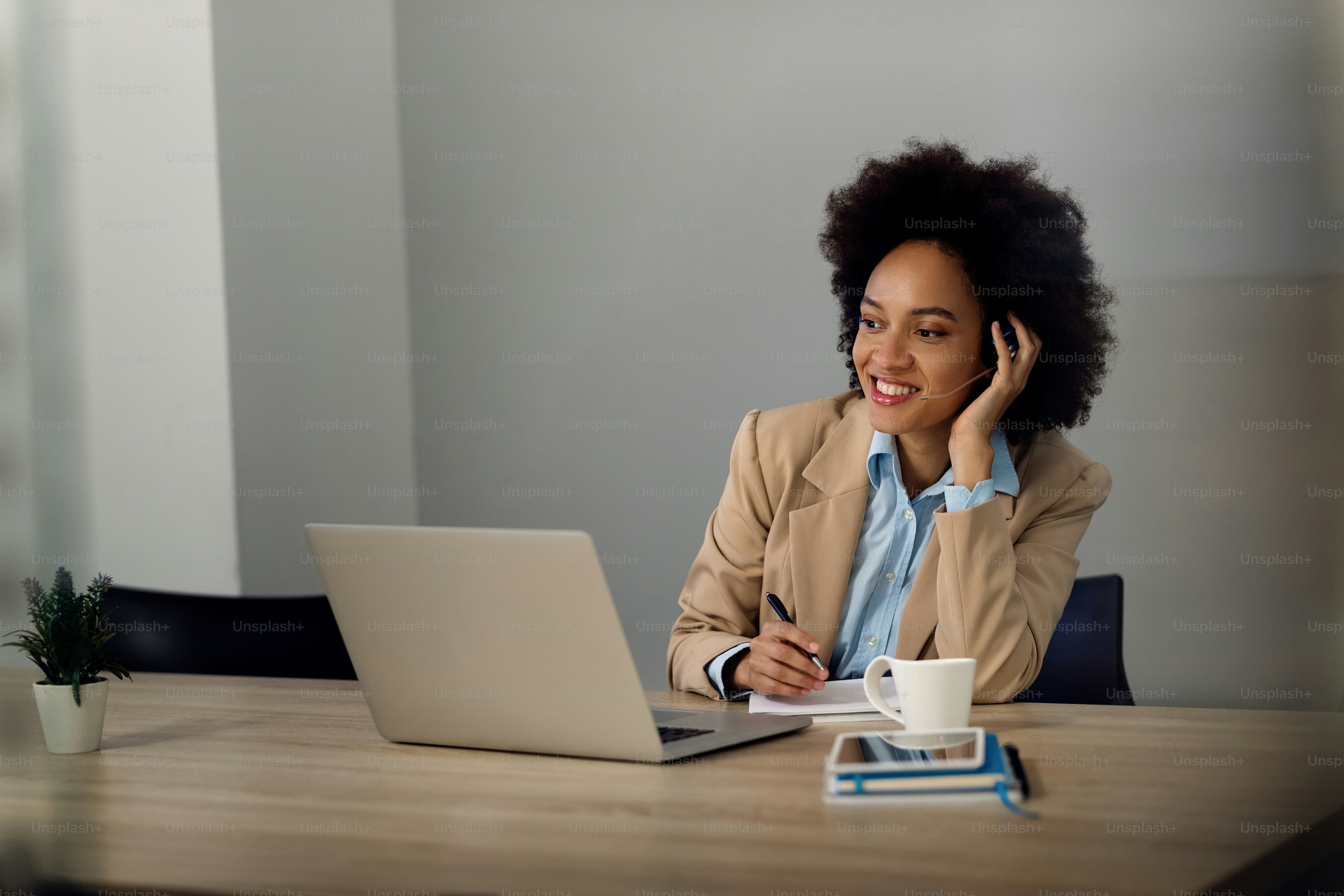 Happy African American businesswoman using computer while making conference call and writing notes in the office.