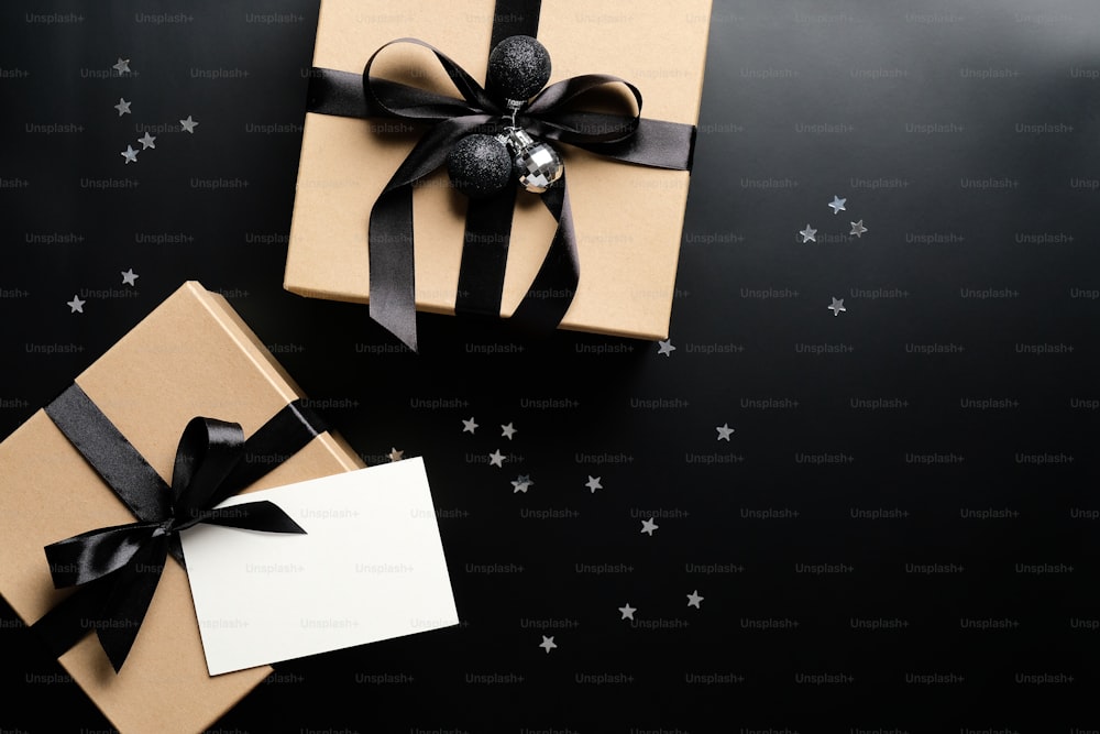 Weihnachtsgeschenke mit Bandschleife und leerer Papierkarte auf schwarzem Grund mit Konfetti. Weihnachtsgeschenke, saisonale Feiertage Grüße Konzept.