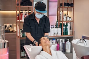 Stylish hairdresser in protective mask performing hair care for the female client in beauty salon