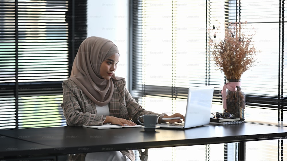 A muslim woman in headscarf is working on her project with computer laptop in office.