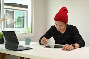 A graphic designer or photographer in red wool hat hand is holding stylus pen drawing on digitizer.