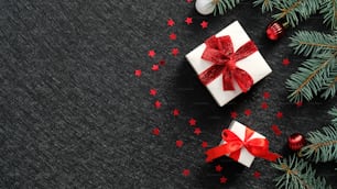 赤いリボンの弓、モミの木の枝、黒い背景にきらめく紙吹雪とエレガントなクリスマスギフトボックス。クリスマスバナーデザイン、新年のグリーティングカードのモックアップ、ヘッダーテンプレート。