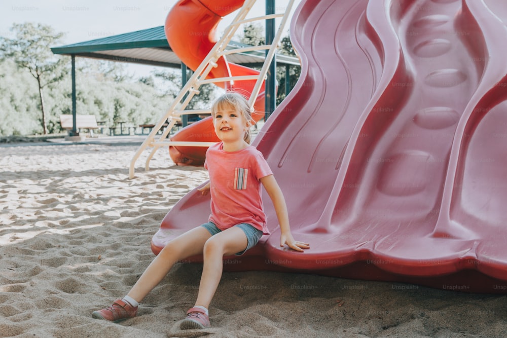 Aktiv glücklich lustig lächelnd kaukasisches Mädchen rutscht auf Spielplatz Schulhof im Freien am Sommer sonnigen Tag. Kind hat Spaß. Saisonale Kinderaktivität draußen. Authentisches Lifestyle-Konzept der Kindheit.