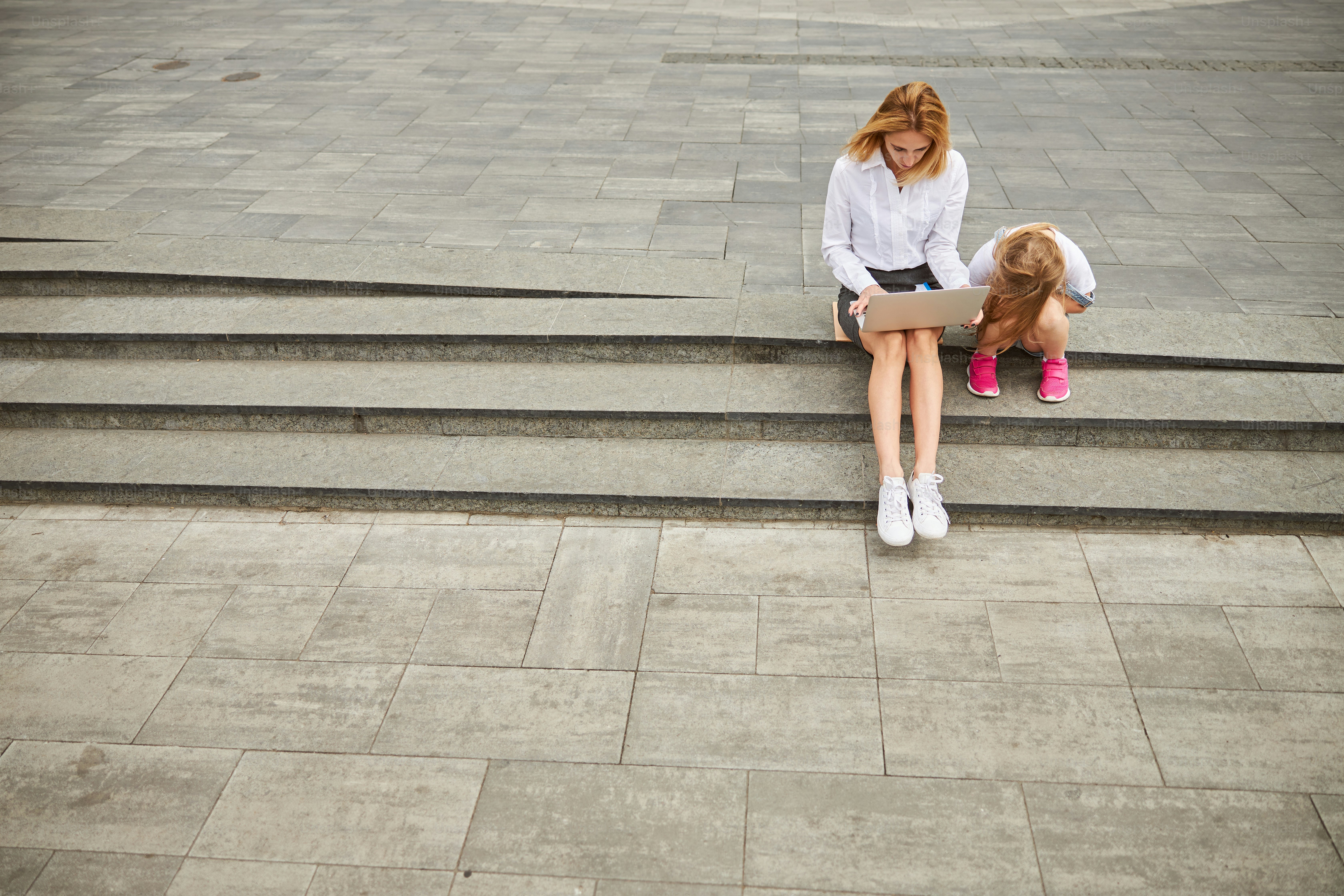 Adult beautiful lady spending time in the outdoors at the laptop while sitting on the steps with her little female child