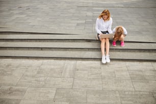 Adult beautiful lady spending time in the outdoors at the laptop while sitting on the steps with her little female child