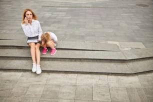 Ganzkörperporträt einer Geschäftsfrau, die mit ihrer Tochter auf den Stufen sitzt, während sie auf dem Handy spricht