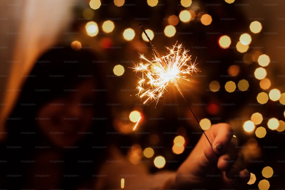 Young female celebrating new year with firework. Happy New Year! Stylish happy woman holding burning sparkler on background of  christmas tree with lights in dark festive room