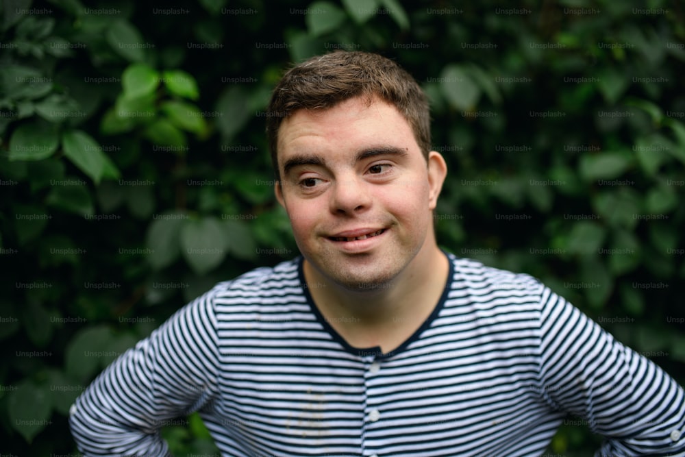 Portrait of down syndrome adult man standing outdoors at green leaves background.