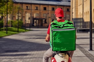 Rear view of a male courier with thermo bag riding bicycle along beautiful sunny street. Food delivery concept