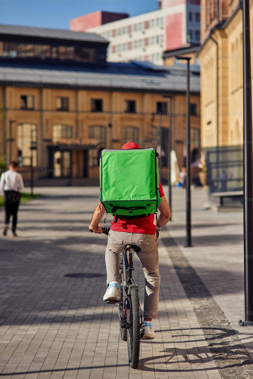Vista trasera de un mensajero con bolsa térmica montando bicicleta a lo largo de una calle de baldosas en un día soleado, tiro vertical de longitud completa. Concepto de entrega de alimentos