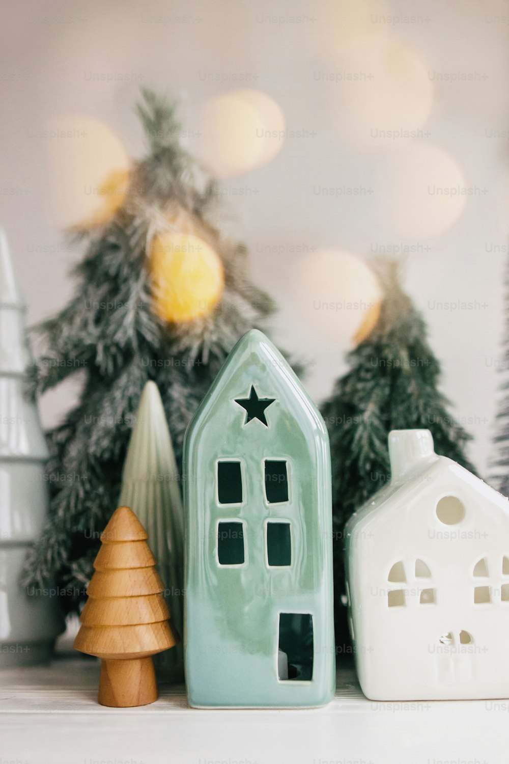 Christmas scene, miniature holiday village. Christmas lights, little ceramic houses, wooden and snowy fir trees on white background. Festive modern decorations on table. Merry Christmas