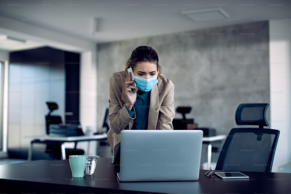 Junge Geschäftsfrau, die während der COVID-19-Pandemie eine E-Mail auf einem Computer liest und telefoniert, während sie im Büro arbeitet.