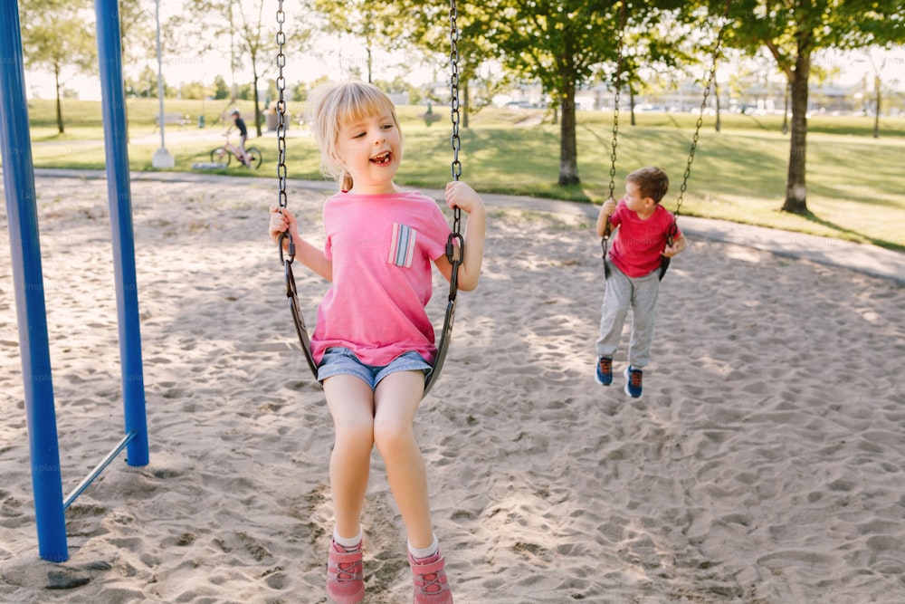 Happy smiling little preschool girl and boy friends swinging on swings at playground outside on summer day. Happy childhood lifestyle concept. Seasonal outdoor activity for kids.