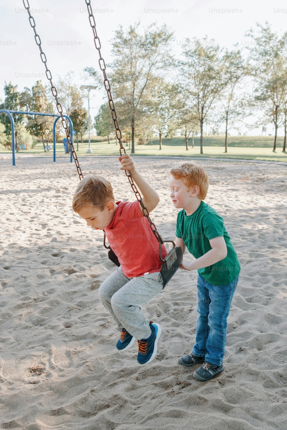 Happy smiling little preschool boys friends swinging on swings at playground outside on summer day. Happy childhood lifestyle concept. Seasonal outdoor activity for kids.