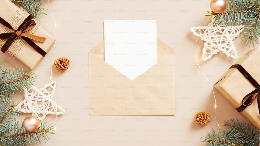 パステルベージュの背景にモミの枝、クリスマスの飾り、ギフトボックスと空白の白いカードのモックアップとクラフト紙の封筒の手紙。クリスマスレターのコンセプト